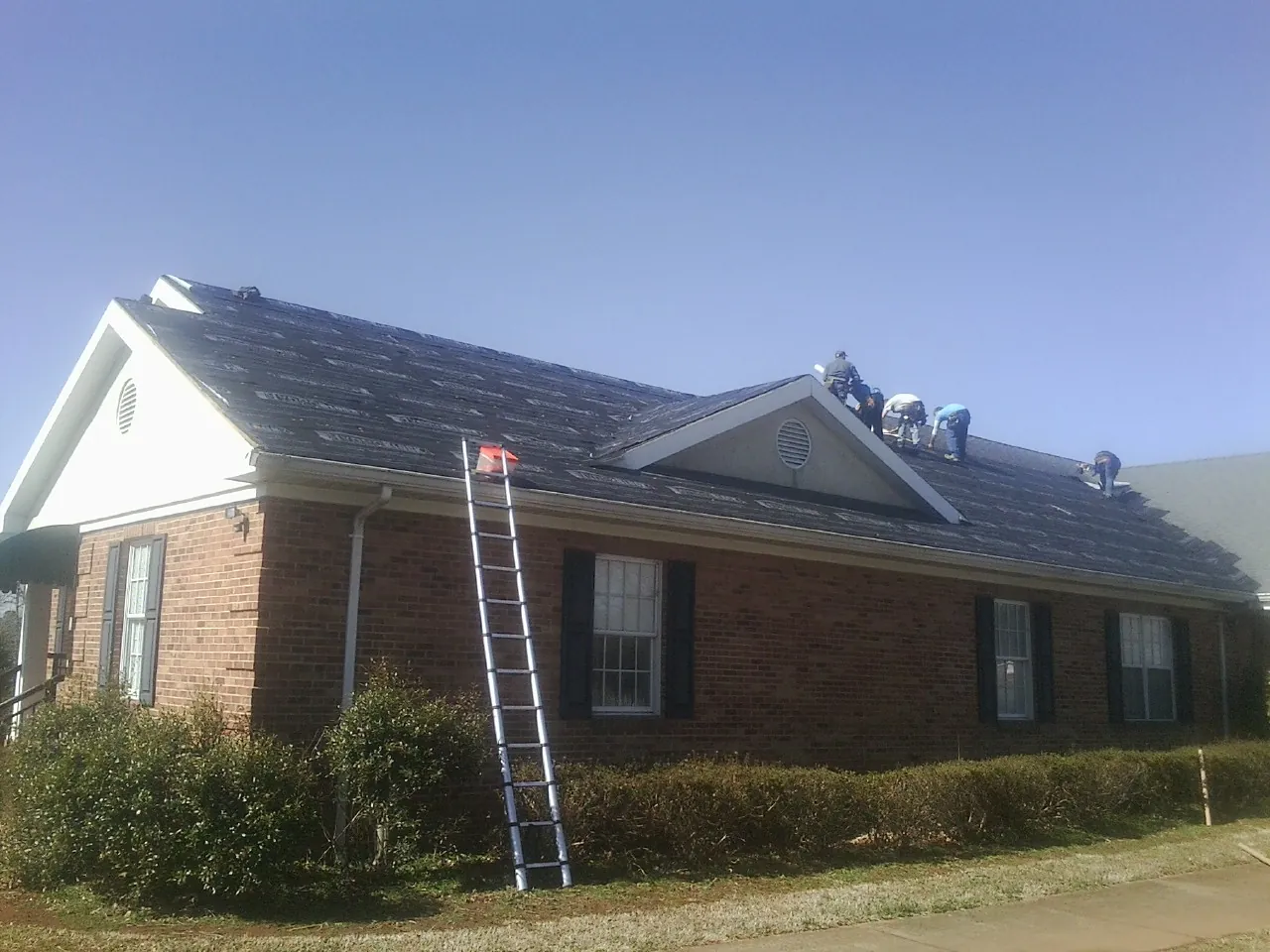 Crew tearing shingles off a brick ranch roof with a ladder set — Emergency Roof Repair work by our crew in Brainard